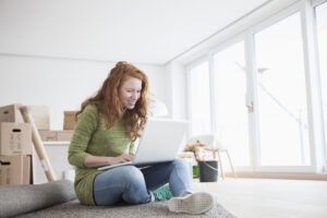 Young woman in new house with cardboard boxes using laptop